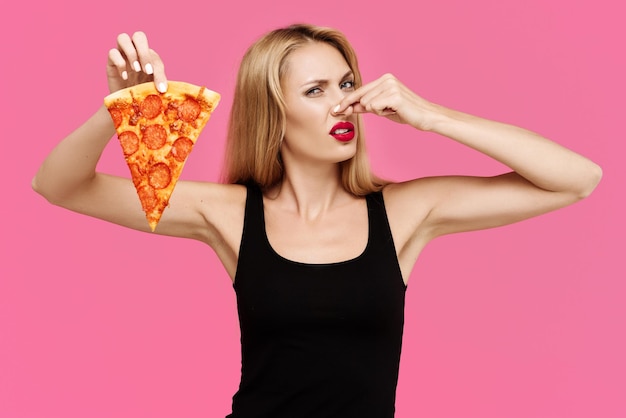 Young beautiful slender girl with a pink background holds pizza in her hands and closes her nose wit