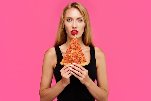 분홍색 배경을 가진 젊고 날씬한 소녀는 건강에 해로운 개념으로 피자를 손에 들고 있습니다