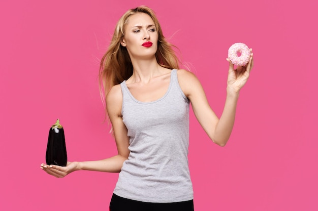 Photo young beautiful slender girl with a pink background with a beautiful smile holding and looking to eggplant and donat concept of un healthy fat junk and healthy food