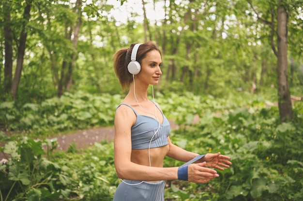 Молодая красивая стройная девушка в синей спортивной одежде с большими белыми наушниками делает упражнение с тренировочной резинкой в парке