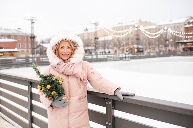 Young beautiful smiling blonde woman wearing coat standing over Christmas city decorations and glow lights hold xmas tree outdoor at ice rink Winter greeting holiday season