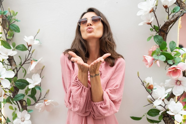 Young beautiful smiling cute romantic woman in trendy summer pink dress Carefree woman posing in the street near white wall with flowers Positive model outdoors in sunglasses