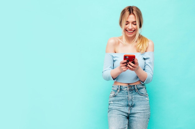 Young beautiful smiling female in trendy summer clothes Sexy carefree woman posing near blue wall in studio Positive brunette model looking at cellphone screen Holding smartphone and using apps