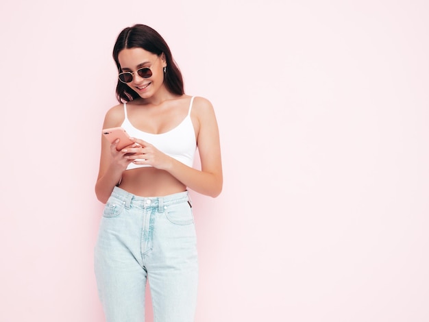 Young beautiful smiling female in trendy summer clothes Sexy carefree woman posing near pink wall in studio Positive brunette model looking at cellphone screen Holding smartphone and using apps