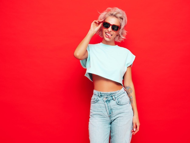 Young beautiful smiling female in trendy summer clothes Sexy carefree woman posing near red wall in studio Positive blond model having fun Cheerful and happy In sunglasses