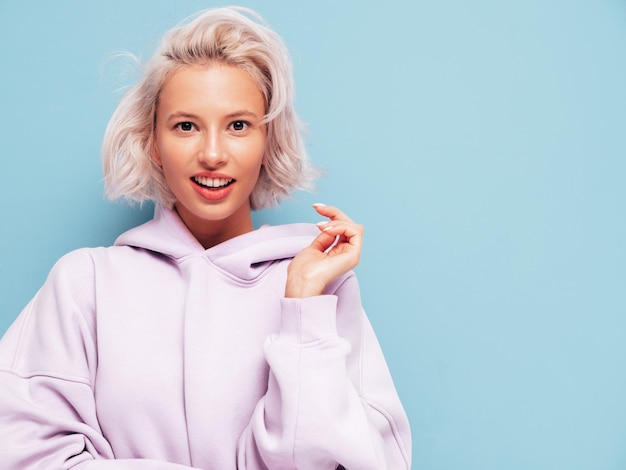 Young beautiful smiling female in trendy summer hoodie Sexy carefree woman posing near blue wall in studio Positive blond model having fun and going crazy Cheerful and happy