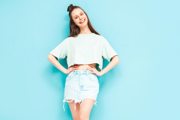 Young beautiful smiling female in trendy summer jeans skirt carefree woman posing near blue wall in studio Positive model having fun indoors Cheerful and happy