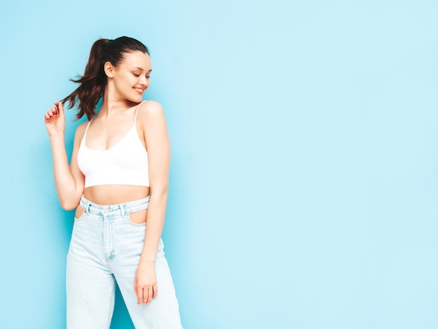 Young beautiful smiling female in trendy summer jeans and top clothes Sexy carefree woman posing near blue wall in studio Positive brunette model having funCheerful and happy