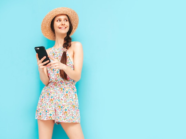 트렌디한 여름 작업복을 입은 젊고 아름다운 미소 스튜디오의 노란색 벽 근처에서 포즈를 취한 섹시한 여성 선글라스와 모자를 쓴 긍정적인 모델 스마트폰 보기 앱 사용