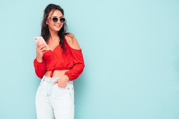 Young beautiful smiling female in trendy summer red top and jeans clothes Sexy carefree woman posing near blue wall in studio Looking at smartphone screen Model using cellphone apps