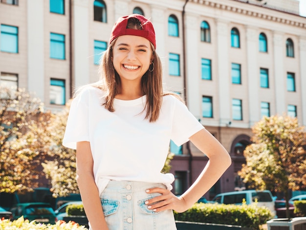 Young beautiful smiling hipster woman in trendy summer white tshirt sexy carefree woman posing on the street background in cap at sunset positive model outdoors cheerful and happy