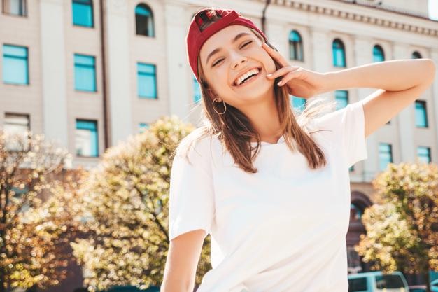 トレンディな夏の白いTシャツで若い美しい笑顔の流行に敏感な女性日没時のキャップで通りの背景にポーズをとるセクシーな屈託のない女性ポジティブモデル屋外陽気で幸せ