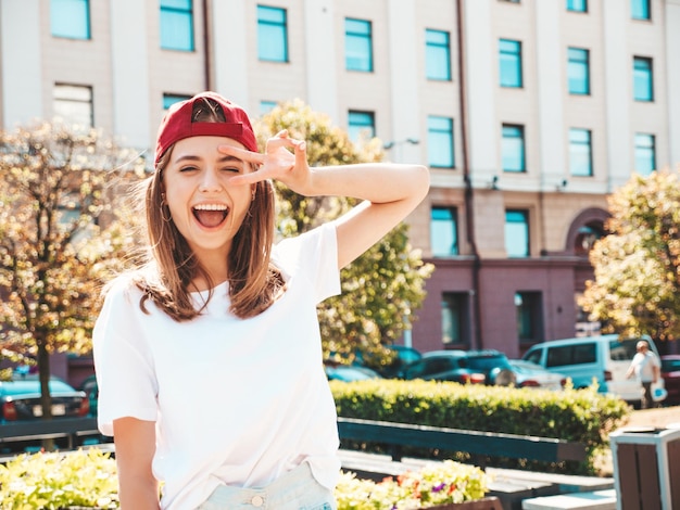 Young beautiful smiling hipster woman in trendy summer white tshirt Sexy carefree woman posing on the street background in cap at sunsetPositive model outdoors Cheerful and happyShows peace sign