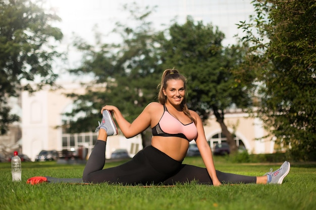 Young beautiful smiling plus size woman in pink sporty top and leggings sitting on twine on yoga mat joyfully looking in camera while stretching in city park