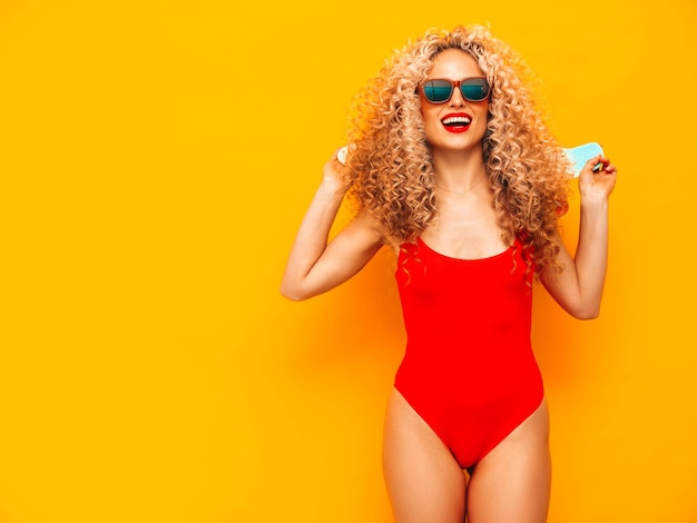 Young beautiful smiling woman posing near yellow wall in studioSexy model in red swimwear bathing suitPositive female with curls hairstyleHappy and cheerful