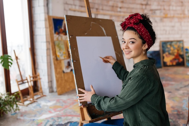 Young beautiful smiling woman with dark curly hair sitting on chair drawing picture on canvas happily looking in camera in big cozy art workshop with big window