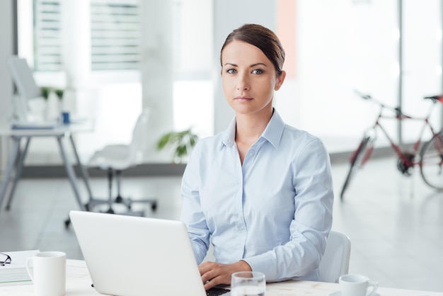 Young beautiful smiling woman working at office desk typing on a laptop and looking at camera