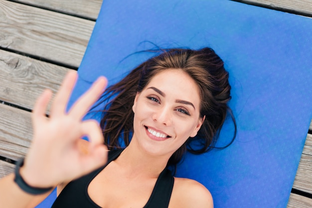 Young beautiful sport woman lies on mat and shows okey symbol.