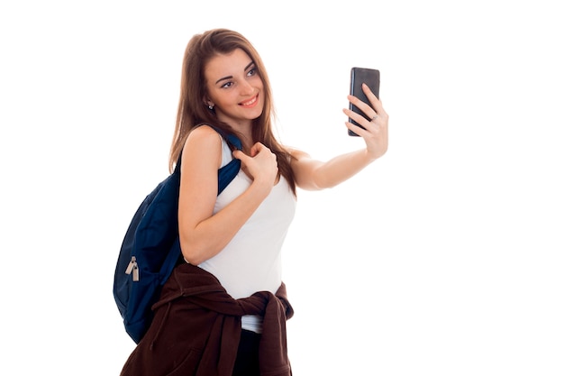 Young beautiful student woman with backpack makes selfie isolated on white wall in studio