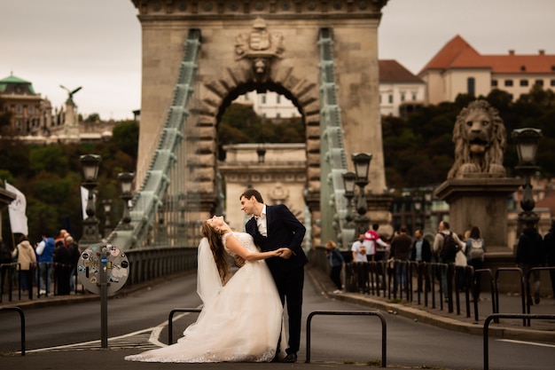 Giovani belle coppie alla moda delle persone appena sposate su un ponte a budapest, ungheria. bella donna in un abito da sposa bianco e bell'uomo in tuta.