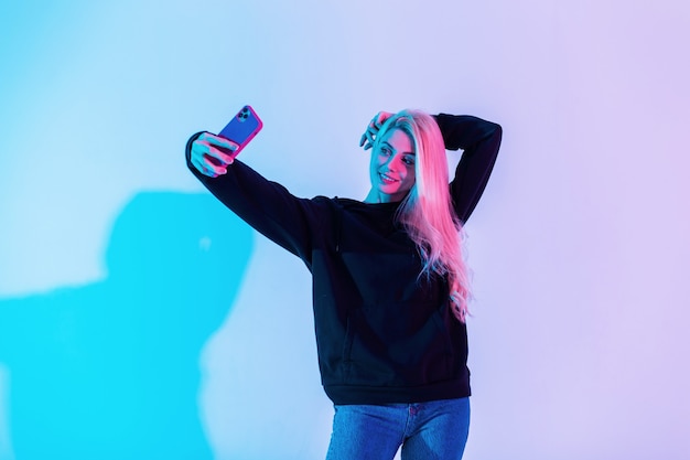 Young beautiful stylish woman model in a fashion black hoodie with blue jeans with a phone makes a selfie picture in a neon pink background in the studio