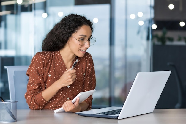 ヒスパニック系の女性のオフィスに座ってリモートで勉強している若い美しく成功したビジネスウーマン