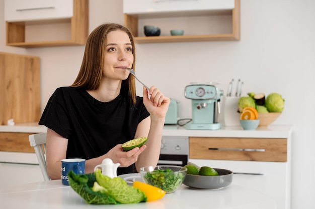 Young beautiful vegetarian girl dressed in pajamas eating fruits and vegetables for breakfast at home in the kitchen