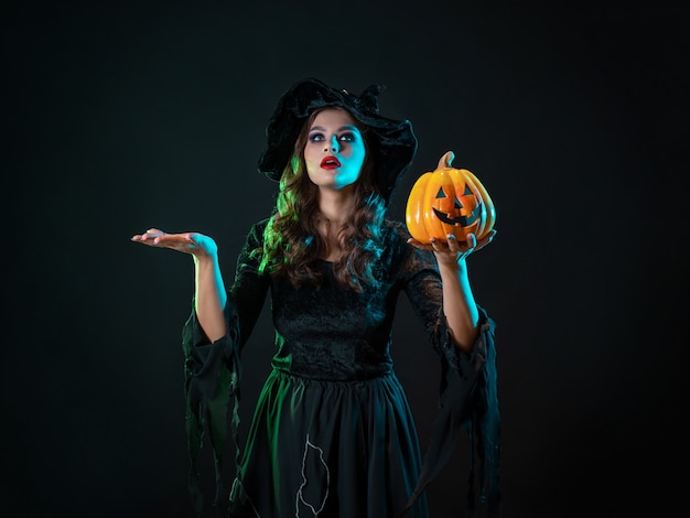 A young beautiful witch in a conical hat holds a halloween pumpkin in her hands