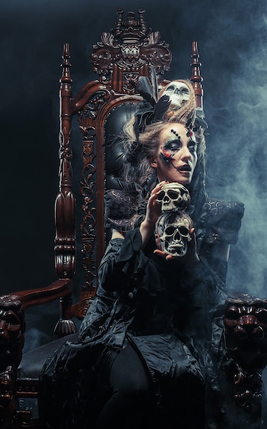 Young beautiful witch sits on a chair. Bright make up, skull, smoke-  halloween theme.