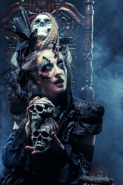 Young beautiful witch sits on a chair. Bright make up, skull, smoke-  halloween theme.