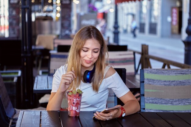 金髪で長い髪の25代の若い美女がストリートカフェに座って、携帯電話を持ってデザートを食べながら笑っている。