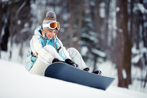 Young beautiful woman adjusts her bindings on snowboard sitting at ski slope