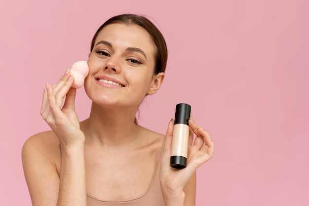 Young beautiful woman applying foundation with sponge and looking at camera on pink background. Perfect skin, makeup concept.