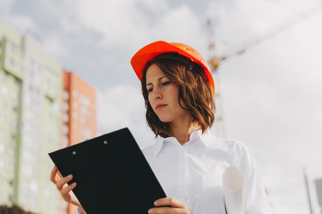 白いシャツとフリップボードを保持しているヘルメットの若い美しい女性建築家。自宅の建設現場でヘルメットをかぶった女性。