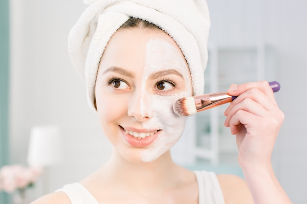 A young beautiful woman in a bathing towel after a shower and cleansing the skin applies a clay mask to the face with a cosmetic brush. beauty, health, cosmetology concept. Lifestyle. Skin care