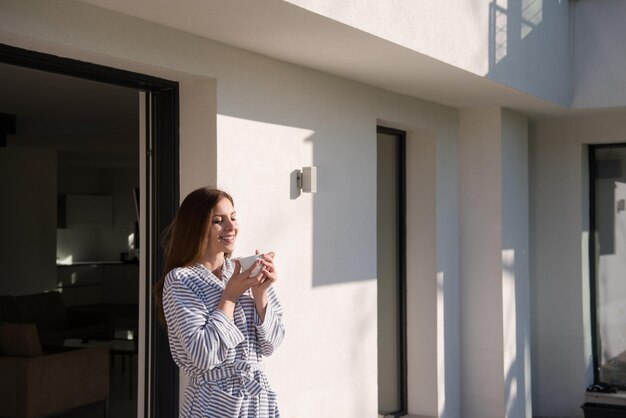 彼女の豪華な家の別荘の前で朝のコーヒーを楽しんでいるバスローブを着た若い美しい女性