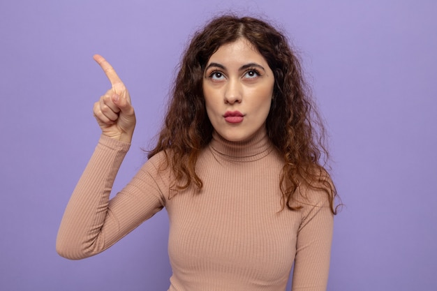 Young beautiful woman in beige turtleneck looking up with serious face pointing with index finger to the side