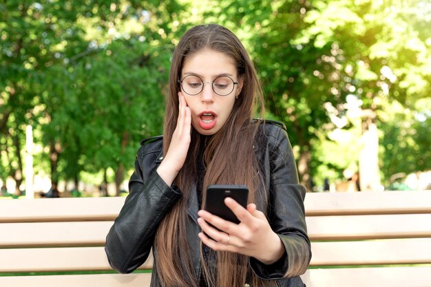 Young beautiful woman in a black leather jacket writes a message or plays games or watches videos or social media news on the phone sitting on a bench in a beautiful green summer or spring park