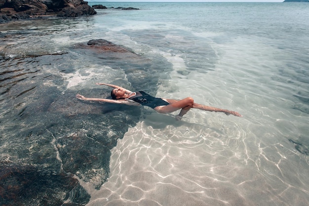 Young beautiful woman in black swimsuit relaxes and sunbathes lying in the water on the beach . The view from the top