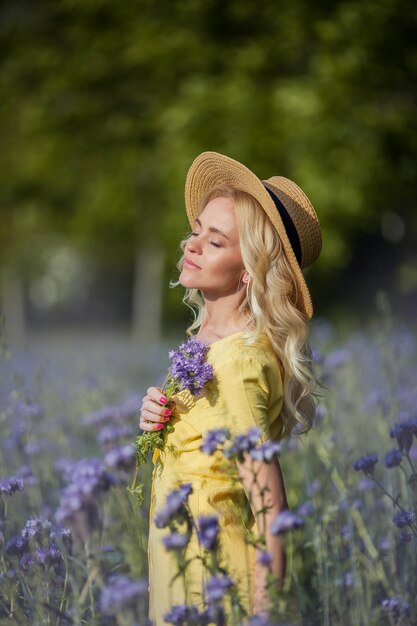모자에 젊은 아름 다운 여자 금발 보라색 꽃의 필드를 안내합니다. 여름. 봄.