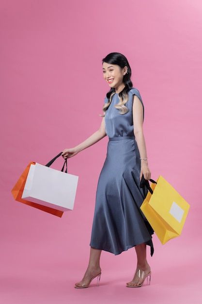 Young beautiful woman in blue dress holding colourful shopping bags isolated over pink background