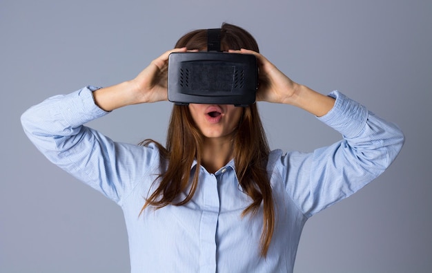 Young beautiful woman in blue shirt using VR glasses showing amazement on grey background in studio