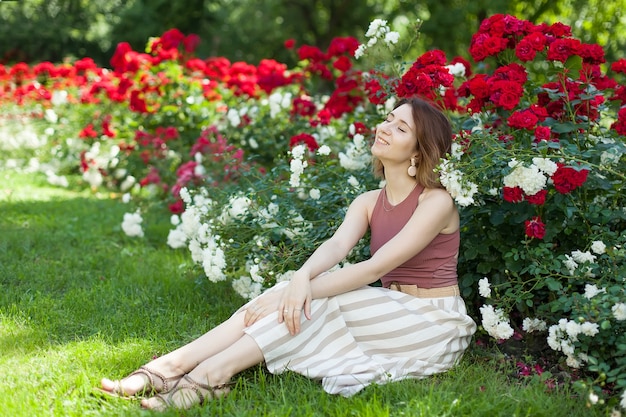 A young beautiful woman in boho clothing is sitting under a Bush of scarlet roses.