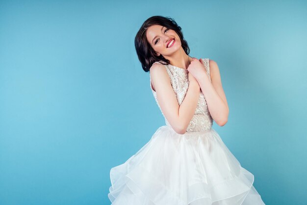 Young and beautiful woman bride with makeup in long chic white wedding dress in studio on a blue background