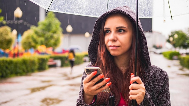 Young beautiful woman browsing mobile phone standing with\
transparent umbrella in rain on city street pretty female using\
smartphone sheltering with umbrella from rain on walk in city\
park