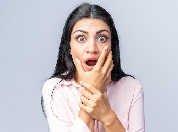 Young beautiful woman in casual clothes  amazed and scared with hand on her face standing over white wall