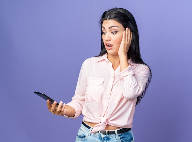 Young beautiful woman in casual clothes holding smartphone looking at it amazed and surprised standing over blue wall