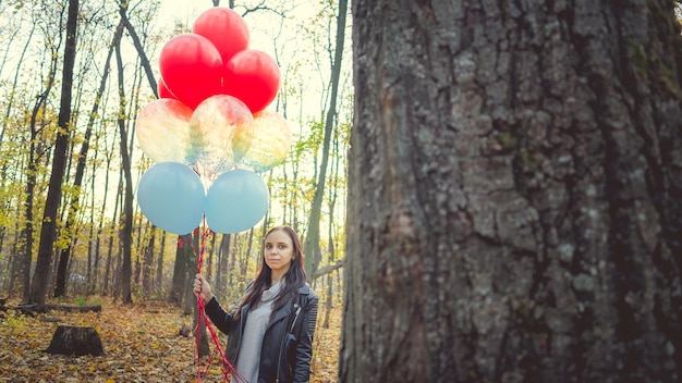 A young beautiful woman in casual clothes is holding the colorful balloons on walk in autumn forest