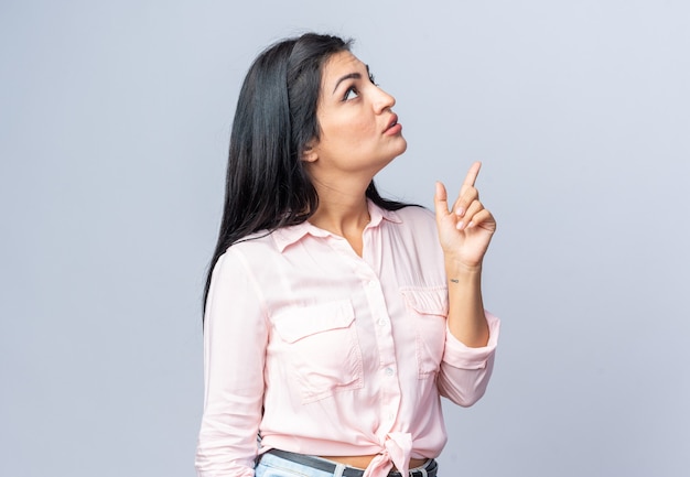Young beautiful woman in casual clothes looking up intrigued pointing with index finger up standing over white wall