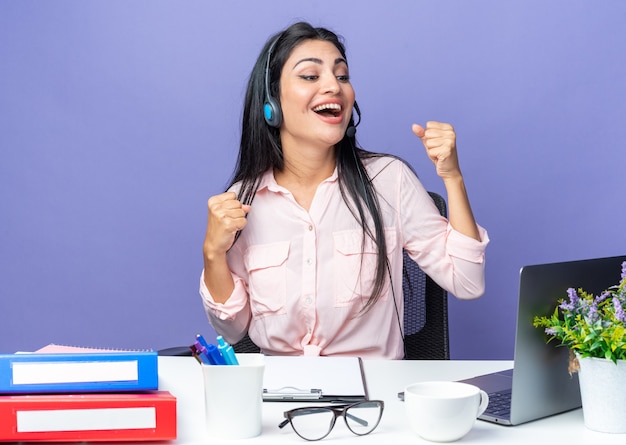 Young beautiful woman in casual clothes wearing headset with microphone happy and excited clenching fists sitting at the table with laptop over blue wall working in office